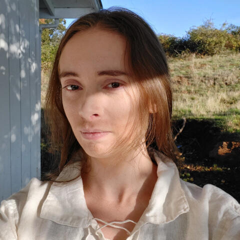 A person with long dark blonde hair smiles slightly at the camera. They have heavy eyebrows and slight stubble and wear a flowing white linen renaissance-style shirt. They are next to a white building in a field.