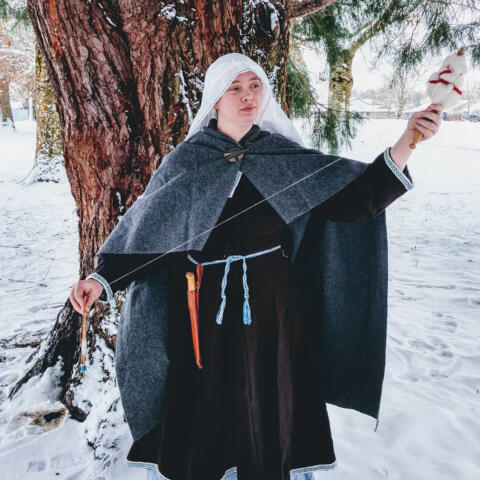 A person with pale skin stands in a snowy field in front of a large cedar tree. They wear a dark overcoat, tablet woven belt with a knife hung from it, a gray cloak and pennannular brooch, and a veil in the style of early Medieval Europe. They hold a short