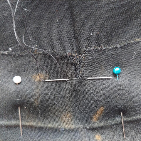 A needle sits in the middle of making a stitch in a trouser hem between two sewing pins. The fabric is old and damaged, with bleach stains.