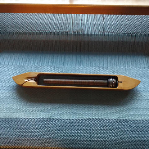 A weaving shuttle with an empty pirn sits on the boundary between gray-blue woven cloth and warp threads on a loom.