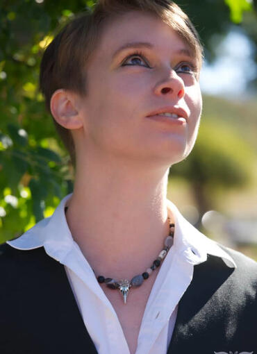 A person with light brown hair and heavy eyeliner and wearing a white collared shirt, black vest, and a beaded necklace looks upward and to the right of the image in the summer sun. Image credit SerenDerryth Creations