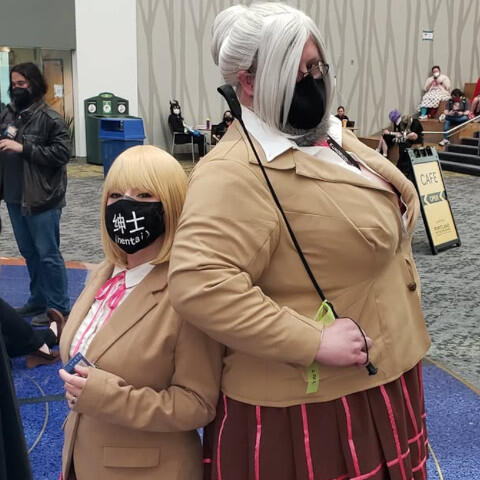 Two cosplayers stand back to back in a convention center. Both wear matching beige blazers and brown pleated skirts with pink ribbon applique stripes. Both are wearing black masks over their noses and mouths. One wears a silver wig styled with bangs and a