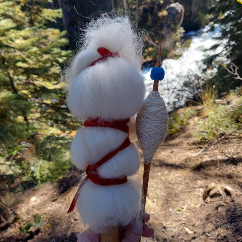 A short distaff dressed with fluffy white wool and a red ribbon is held next to a spindle with a cop of white thread and an ancient blue glass bead. They are held in front of a sunny ravine covered with small coniferous trees and a swift river.