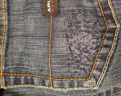 The back pocket of a pair of sturdy denim jeans shows a hole that has been patched and darned.
