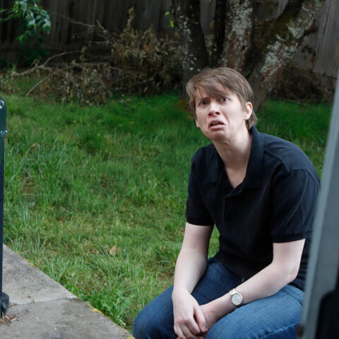 A person with light brown hair wears jeans, a dark blue button-up shirt, and a dismayed expression. They are seated on a patio in a back yard with green grass and a few trees.
